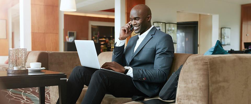 Businessman with beverage using cell phone and laptop in hotel lobby