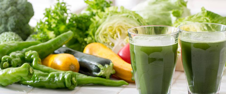 Two glasses of green vegetable juices in front of a display of vegetables