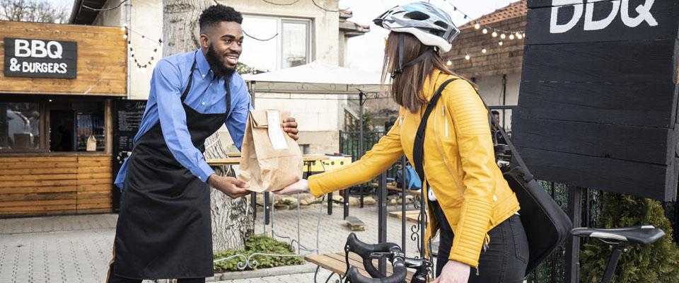 Man wearing apron handing BBQ takeout to a woman on a bicycle