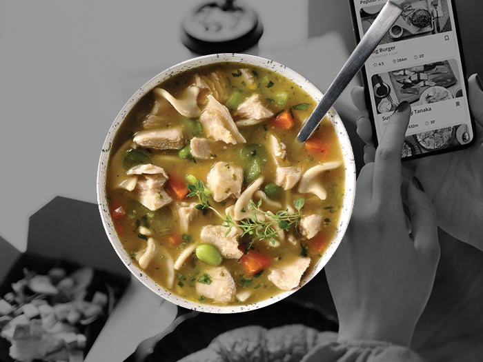 A person enjoys a bowl of Sweet Earth Mindful Chik’n Soup while playing on the cell phone.