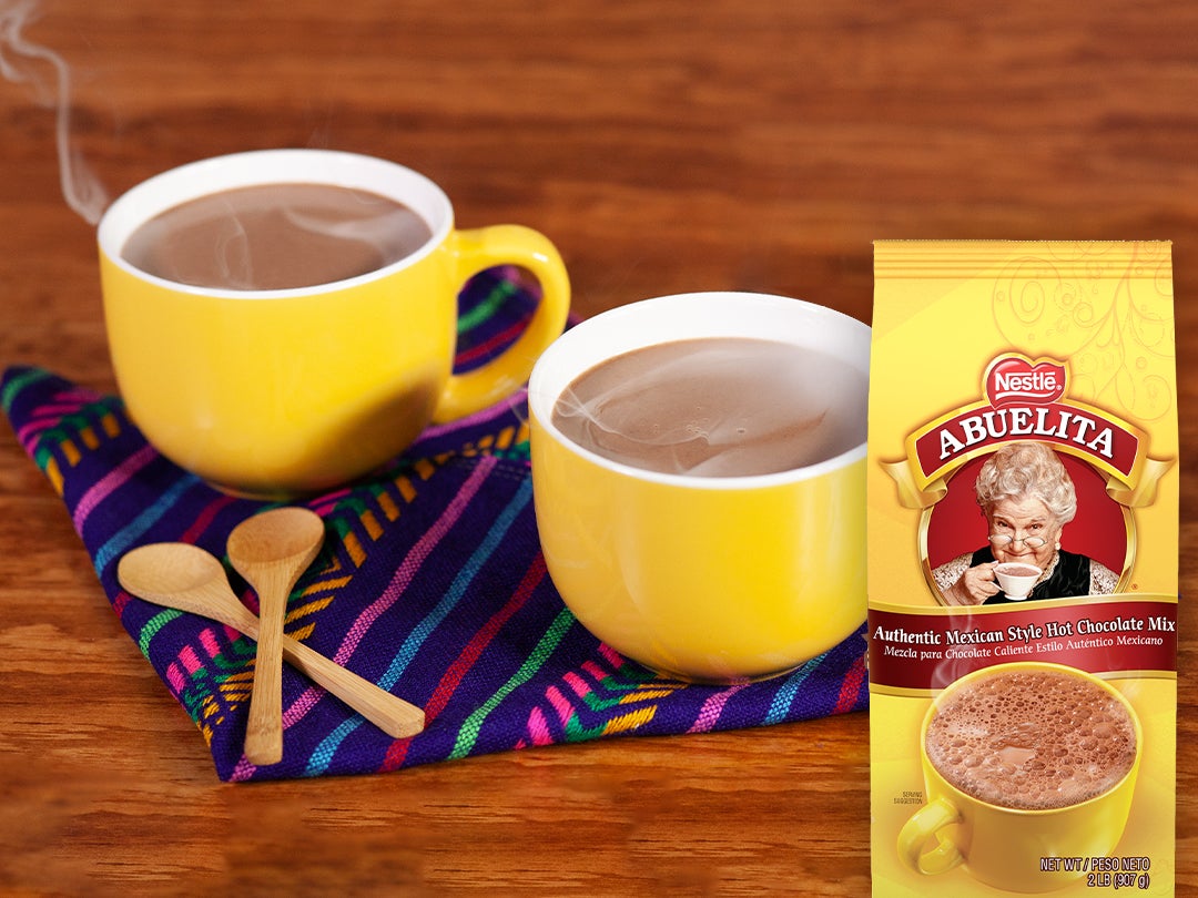 On a wooden surface, two yellow mugs with steaming Mexican-style hot chocolate sit next to two small wooden spoons on a decor