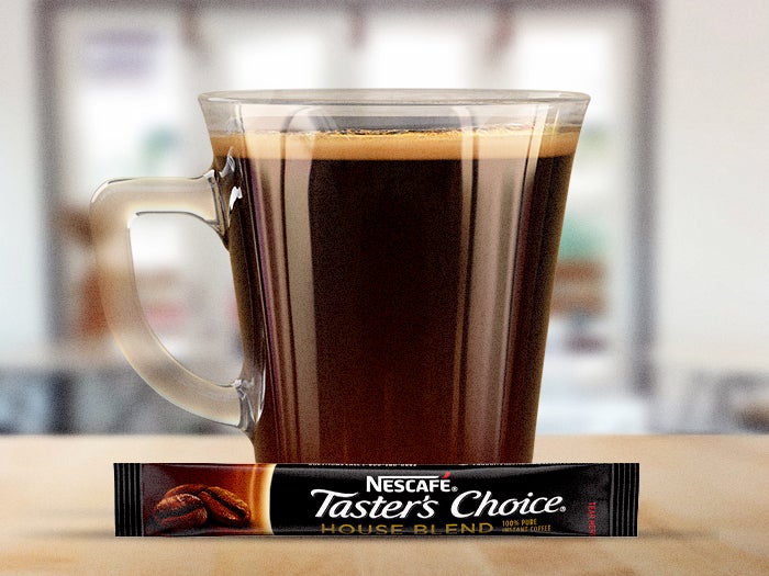 A glass mug filled with Nescafé Taster's Choice House Blend instant coffee, with a single-serve packet on the table.