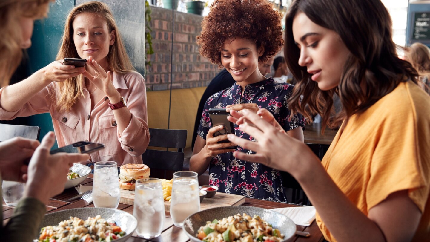 four young adults holding cell phone taking pictures of food