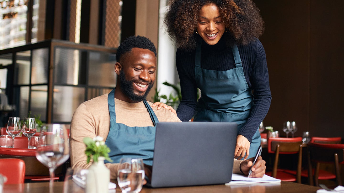 A man in an apron working at a laptop with a woman helping