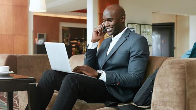 Businessman with beverage using cell phone and laptop in hotel lobby