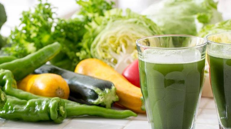 Two glasses of green vegetable juices in front of a display of vegetables