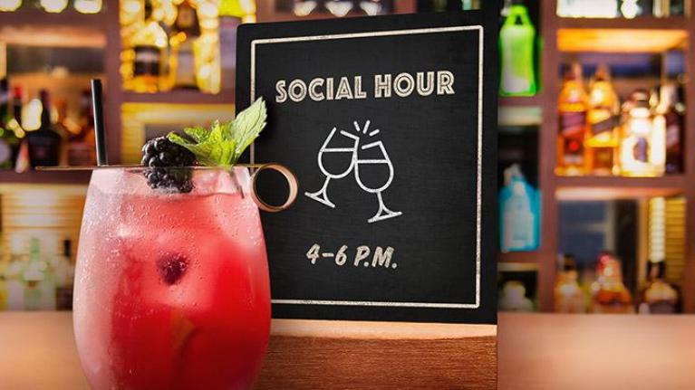 Pink cocktail sitting on bar with social hour sign behind it