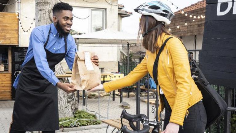 Man wearing apron handing BBQ takeout to a woman on a bicycle