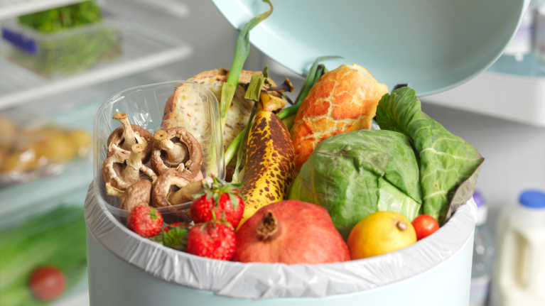 Fruits and vegetables in a white trash can and a refrigerator in the background