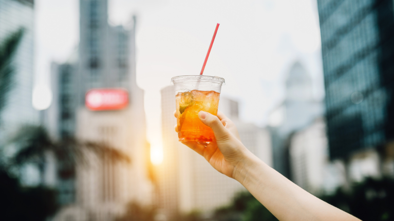Iced tea in a plastic cup with straw being held up high with a city background