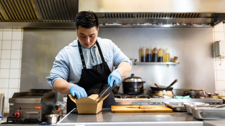 chef in kitchen adding food to a take out container