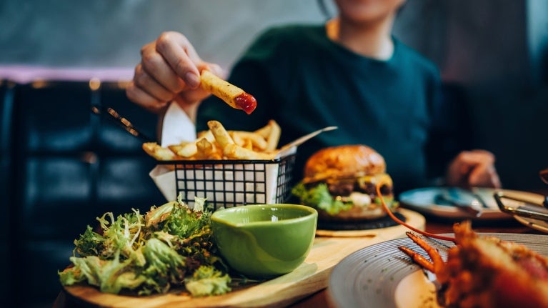 Man holding a french fry dipped in ketchup with broccoli on wooden board