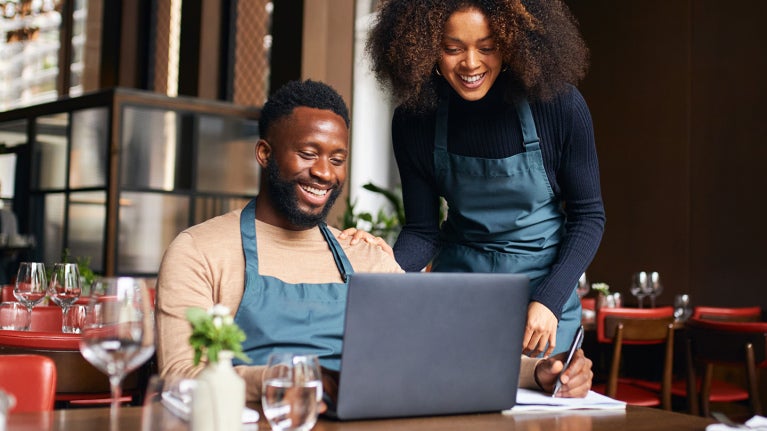 A man in an apron working at a laptop with a woman helping