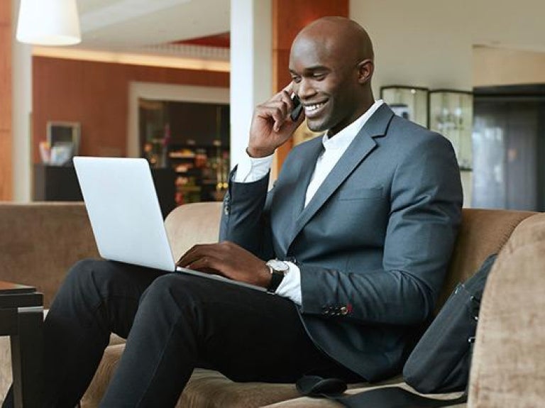 Businessman with beverage using cell phone and laptop in hotel lobby