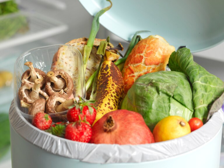 Fruits and vegetables in a white trash can and a refrigerator in the background