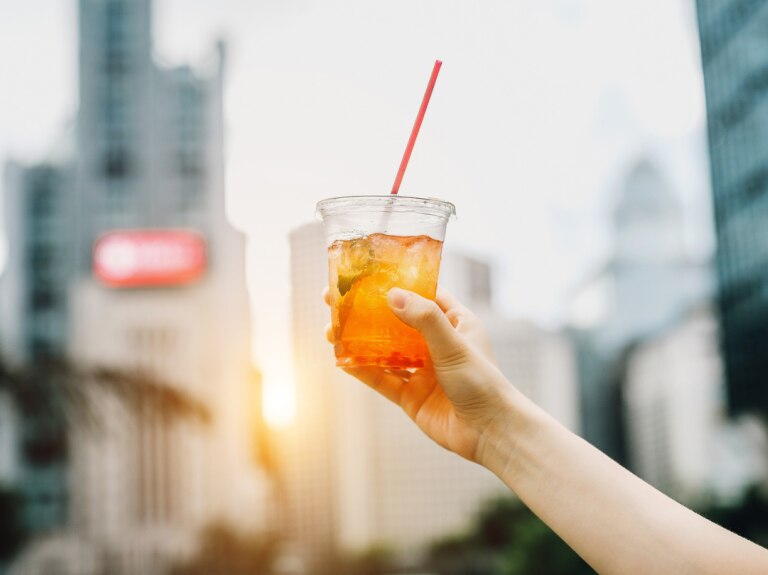 Iced tea in a plastic cup with straw being held up high with a city background