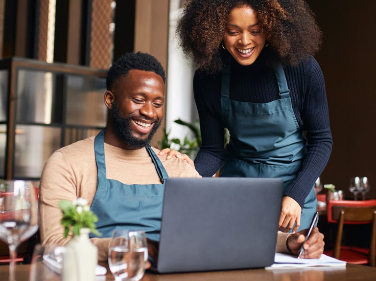 A man in an apron working at a laptop with a woman helping