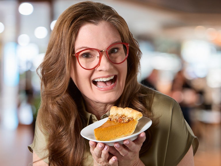 A smiling female consumer proudly holding a white plate with a piece of pumpkin pie.