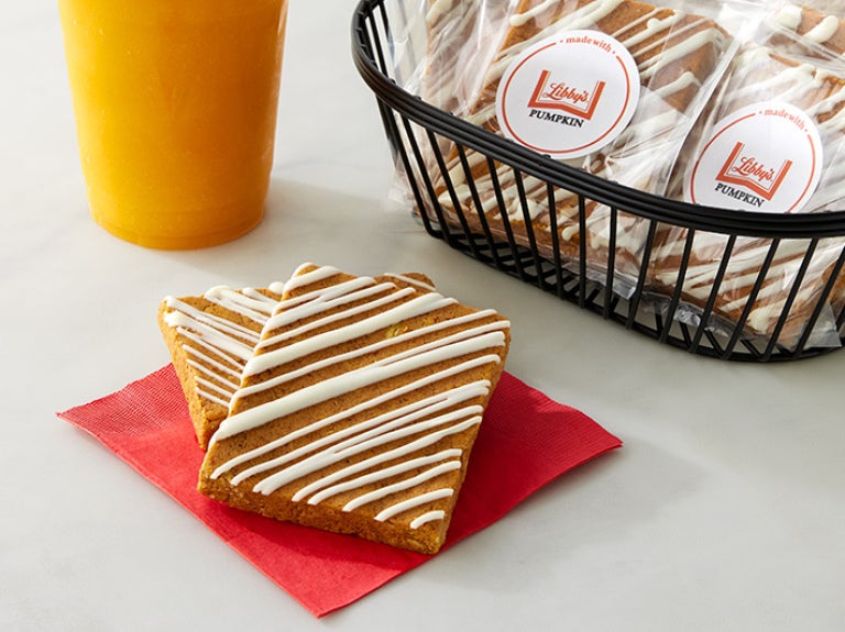 Two frosted pumpkin protein bars with a cup of orange juice and a basket of packaged protein bars in the background.
