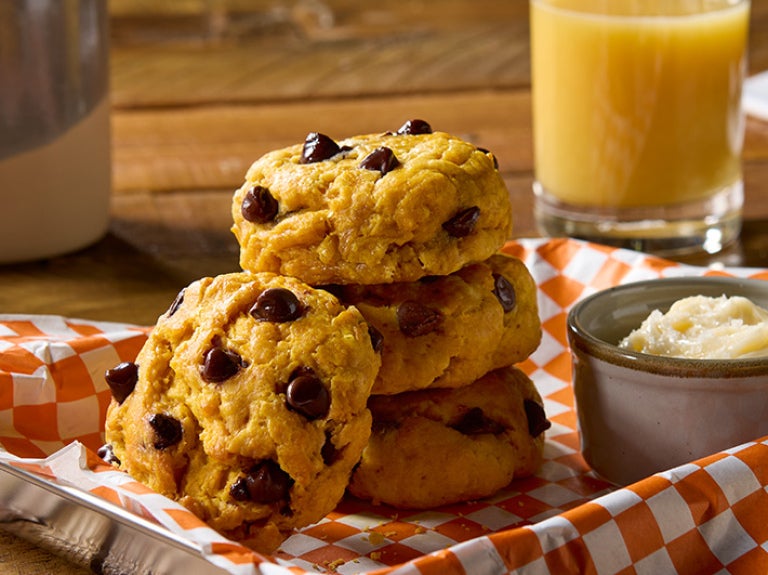 A basket with a stack of pumpkin biscuits filled with chocolate chips and a side of butter and a cup of orange juice.