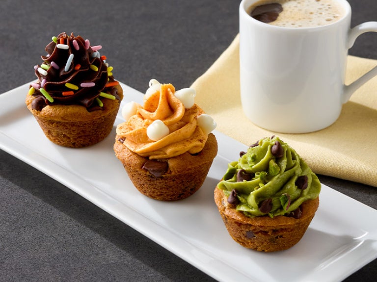 Three pumpkin cookie cups on a rectangular white plate, each with a different frosting crown and sprinkles.