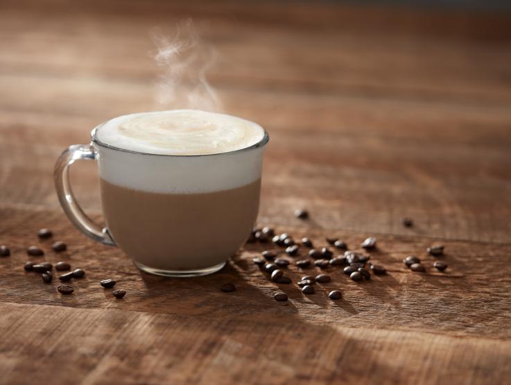 cappucchino steaming in a clear mug and coffee beans scattered around it