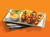 three tacos on a white plate with cheese sauce in a small cup