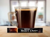 A glass mug filled with Nescafé Taster's Choice House Blend instant coffee, with a single-serve packet on the table.