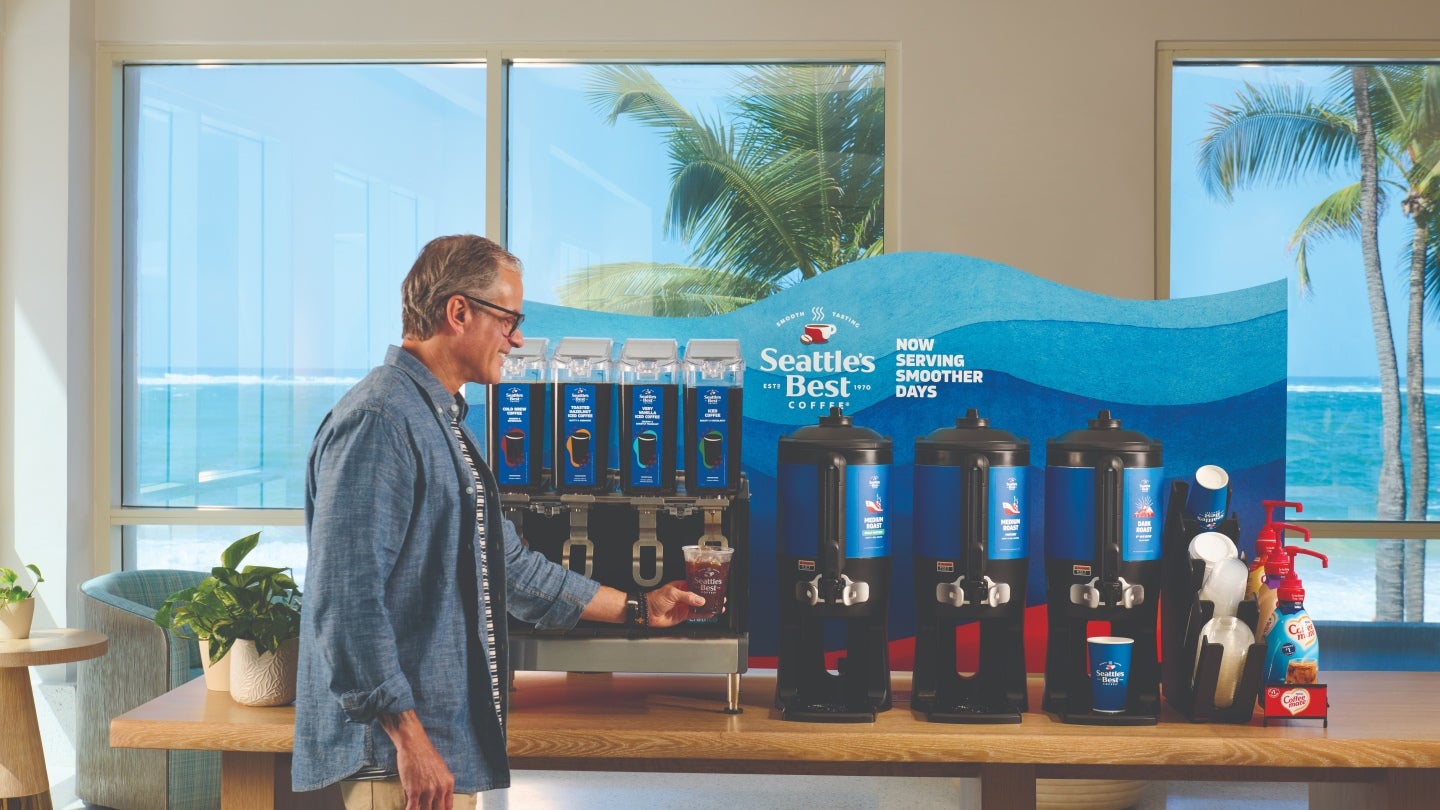 Person serving iced coffee from a self-serve coffee station with tropical beach view.